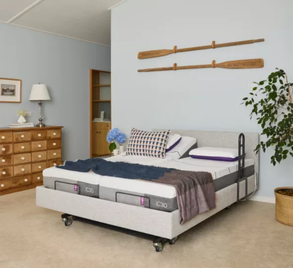 A bed with a light gray upholstered base and black metal side rails, shown in a styled bedroom. The mattress is white with gray side panels branded 'KIKO', and the bed is dressed with blue and purple bedding.