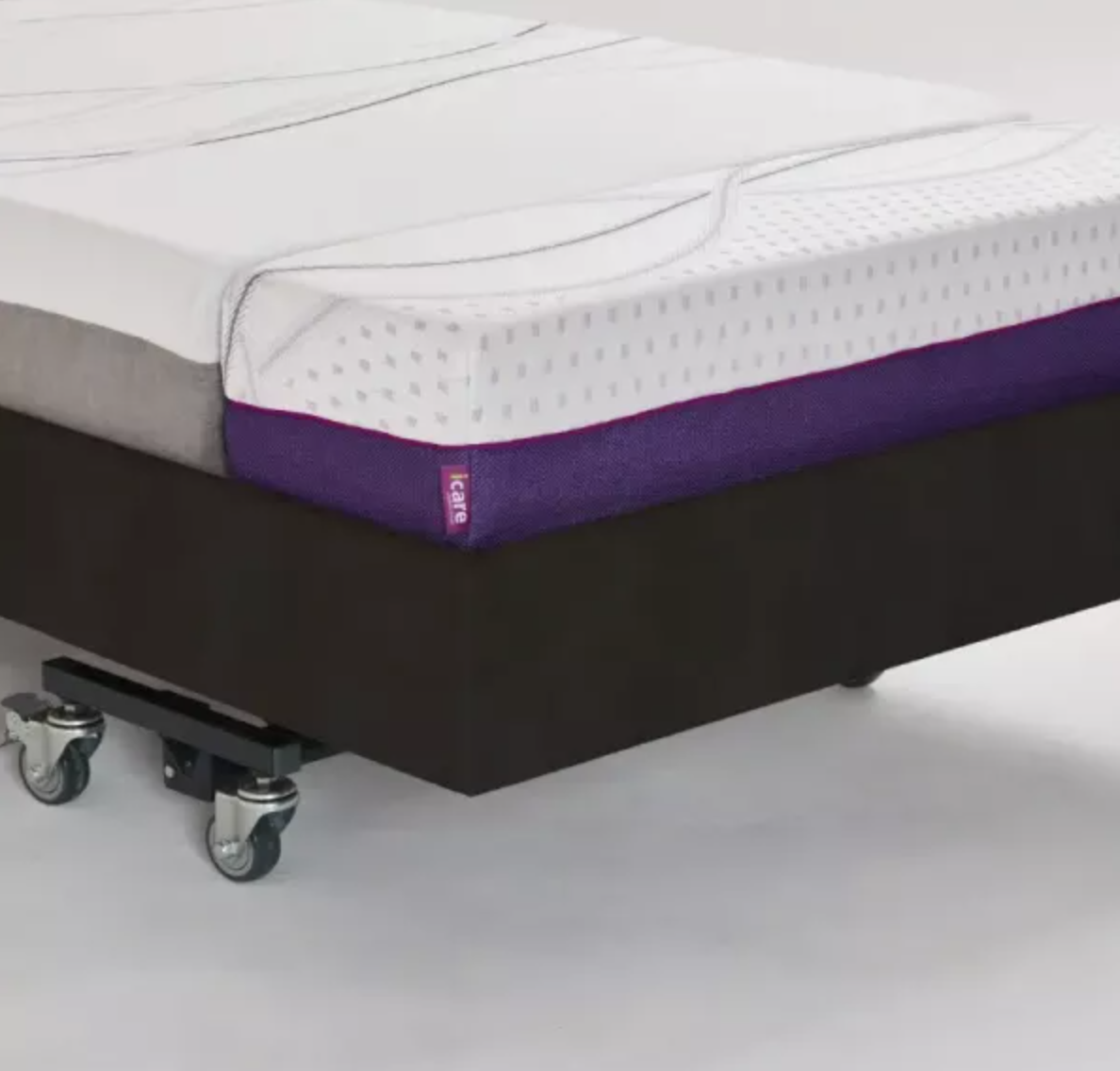 A mattress on a wheeled platform bed frame, shown in a studio setting. The mattress has a white textured top, a purple side bolster, and a gray side panel.
